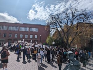 A crowd of students gather as students facing the crowd hold signs at a student union protest in front of a brick building under a blue sky.