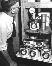 A black and white photo shows a researcher with glasses monitoring cans of Cougar cheese. 