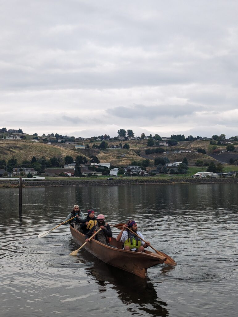 Several people in a hand carved canoe with wood paddles float on the Snake River under a cloudy sky.