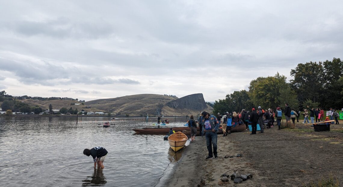 People gather at the shores of the Snake River in Hells Gate State Park outside of Lewiston, Idaho. Desert mountains can be seen in the distance as canoes sit on a sandy shore under clouds.