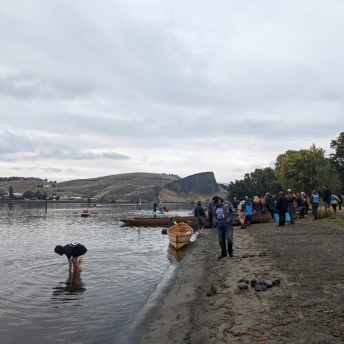 People gather at the shores of the Snake River in Hells Gate State Park outside of Lewiston, Idaho. Desert mountains can be seen in the distance as canoes sit on a sandy shore under clouds.