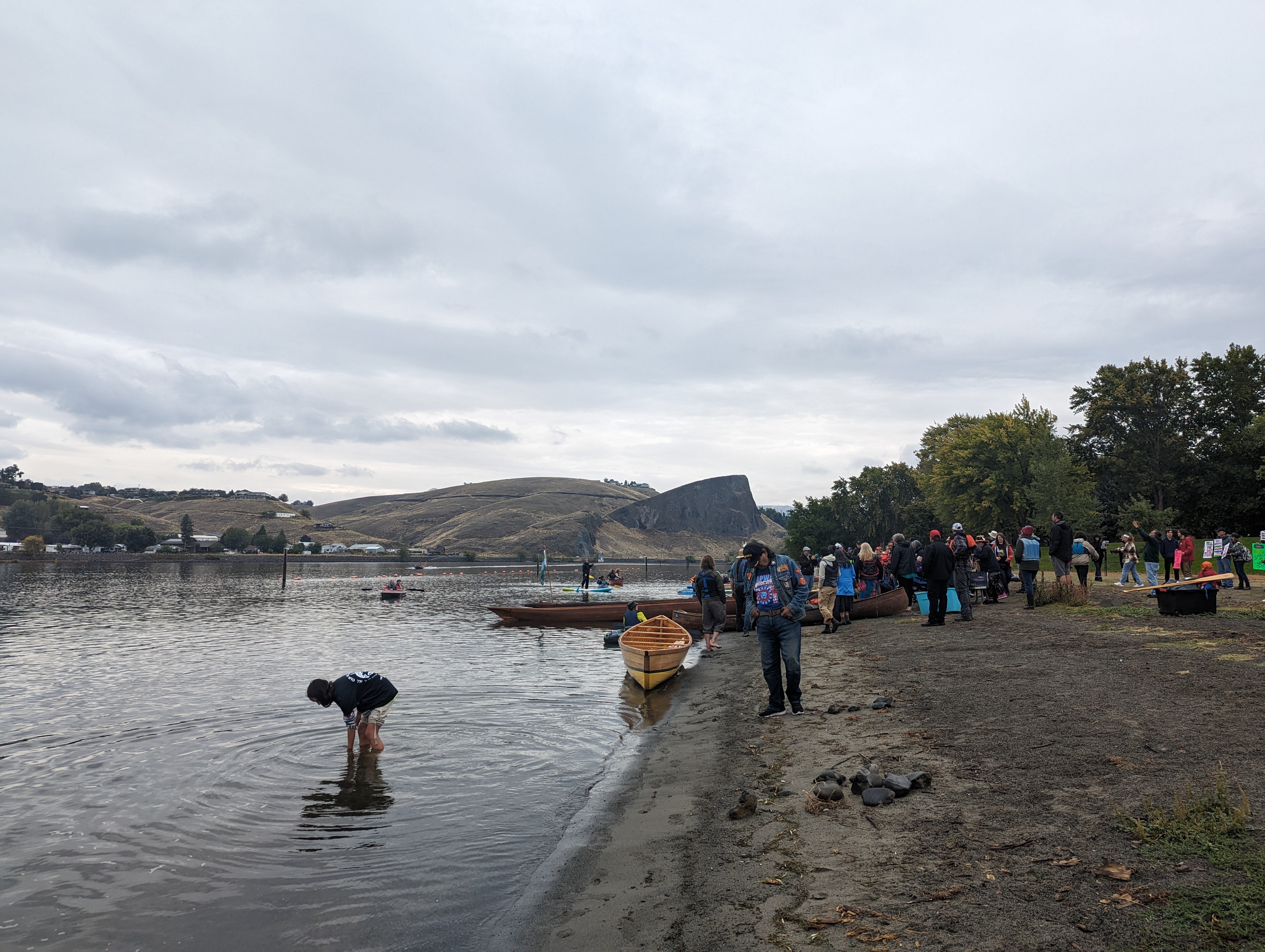 People gather at the shores of the Snake River in Hells Gate State Park outside of Lewiston, Idaho. Desert mountains can be seen in the distance as canoes sit on a sandy shore under clouds.