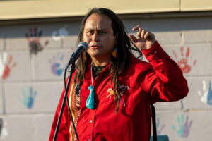 A Native American man with long dark hair in a red shirt stands in front of a brick wall with hand prints painted on it.