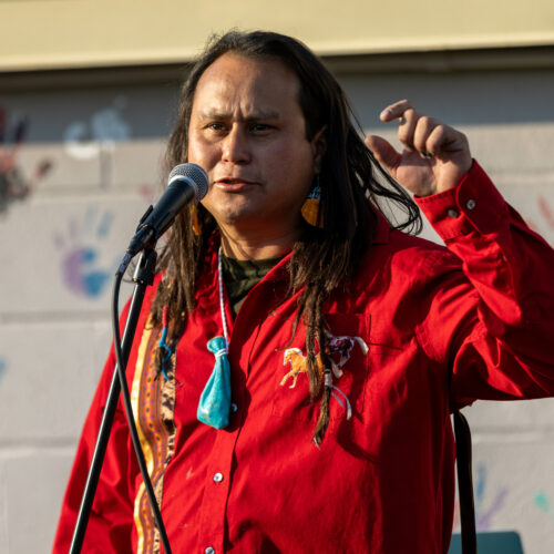 A Native American man with long dark hair in a red shirt stands in front of a brick wall with hand prints painted on it.