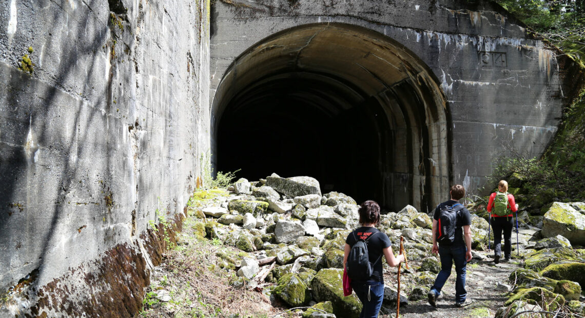 A group of hikers wearing black are standing on a gravel trail next to a pile of gray rocks. They are heading into a dark cement tunnel.