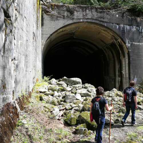 A group of hikers wearing black are standing on a gravel trail next to a pile of gray rocks. They are heading into a dark cement tunnel.