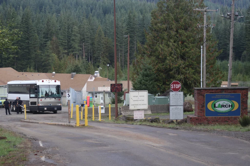 A bus is seen driving out of a parking lot entrance with stop and warning signs around the entrance. A sign mounted to a brick wall on the right side of the road reads "Larch" in big letters. In the background, the white buildings of the prison are visible at the base of an evergreen forest.