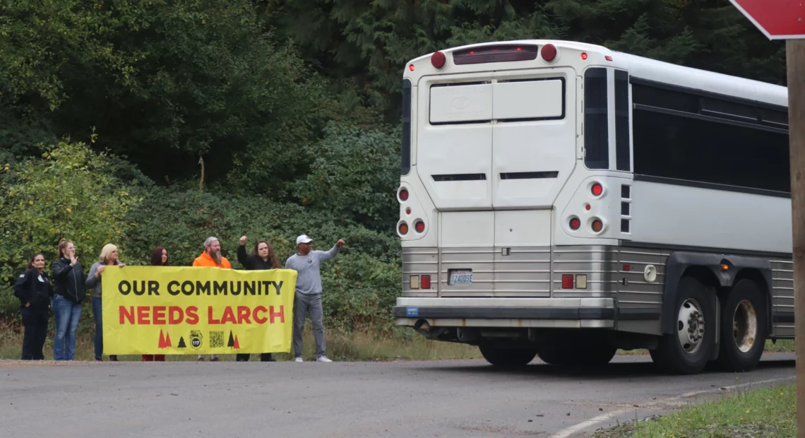A handful of people stand holding a bright yellow banner that reads "Our Community Needs Larch" as the back of a bus is seen driving past them.