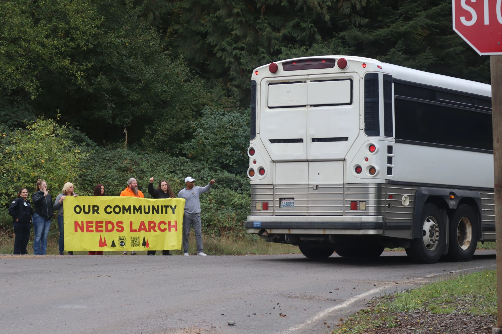 A handful of people stand holding a bright yellow banner that reads "Our Community Needs Larch" as the back of a bus is seen driving past them.