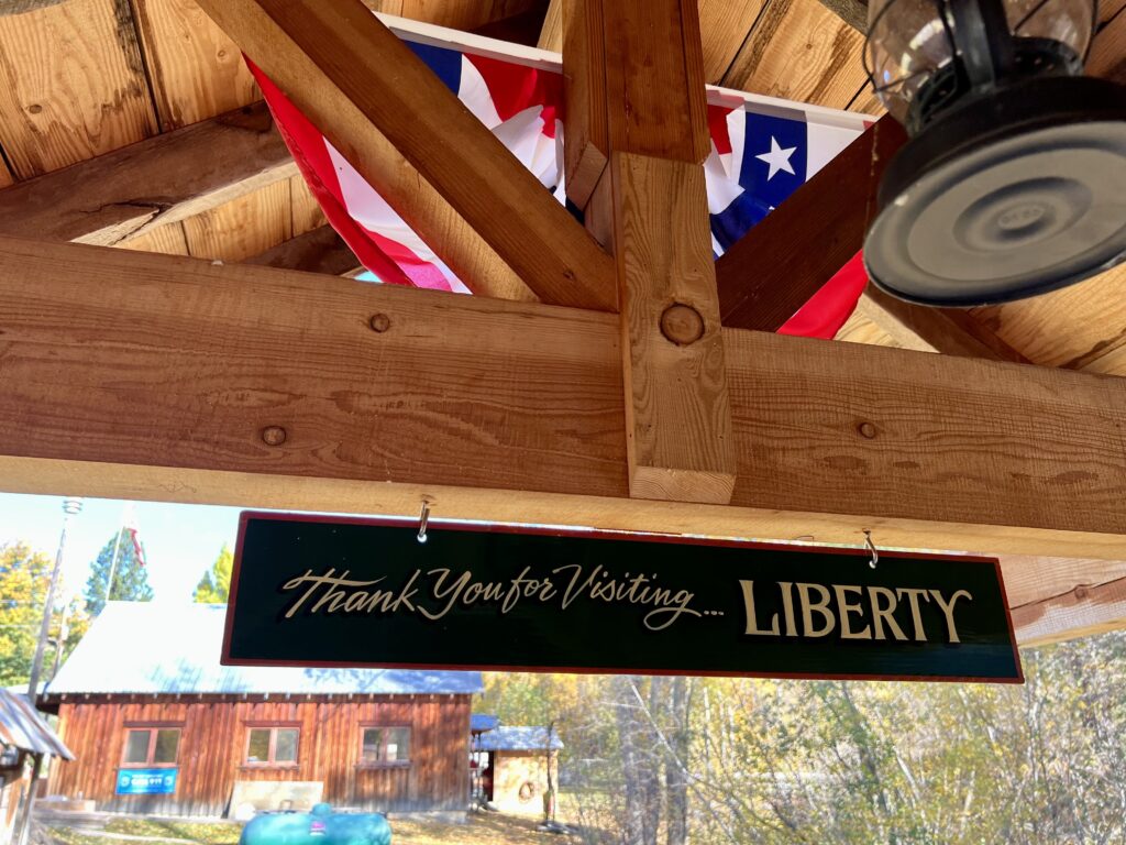 A brown sign that says "Thank you for visiting ... Liberty" hangs on a wooden beam. The beam also has a red, white and blue banner on it.