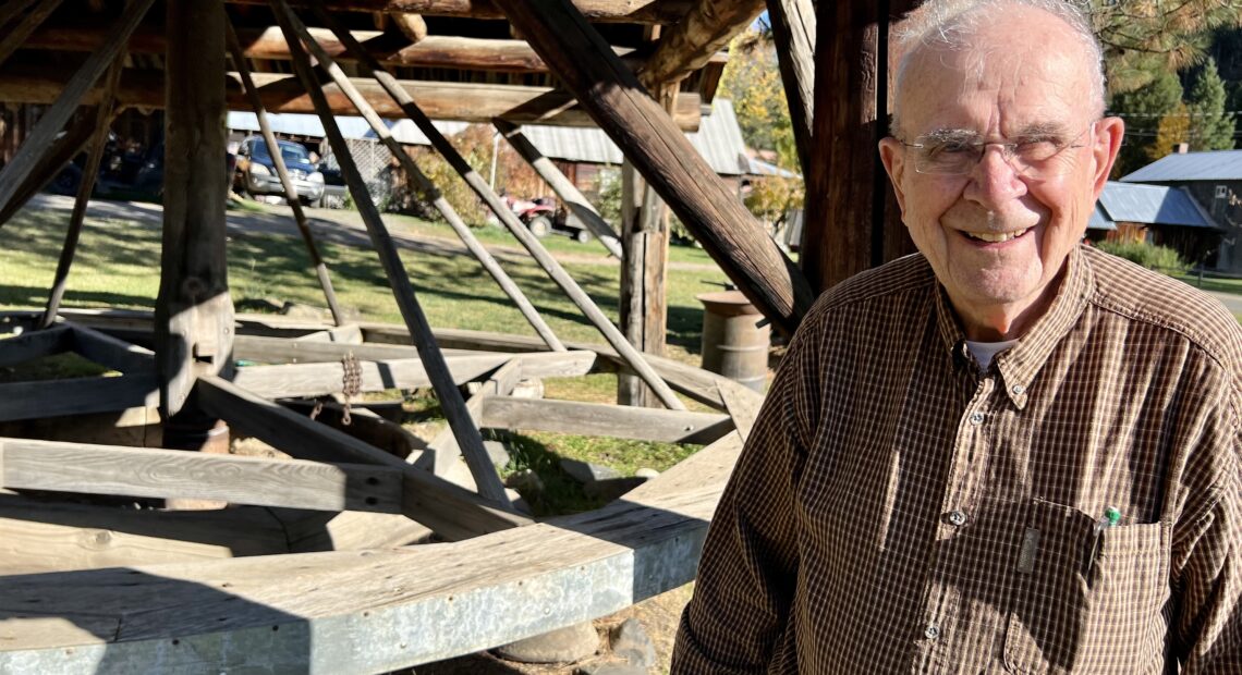 A man wearing a checked button down shirt and glasses is standing in front of what looks like a large wooden carousel.