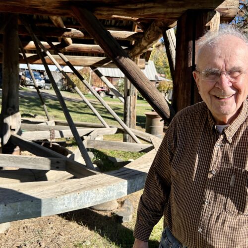 A man wearing a checked button down shirt and glasses is standing in front of what looks like a large wooden carousel.