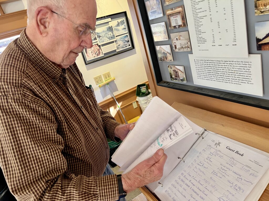 A man in a checkered button down shirt stands in a room with yellow walls. On a wood table is a white guest book with signatures. He is flipping through the guest book