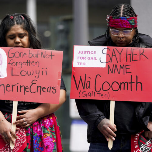 Two Native American protestors hold red signs with the names of Indigenous victims.
