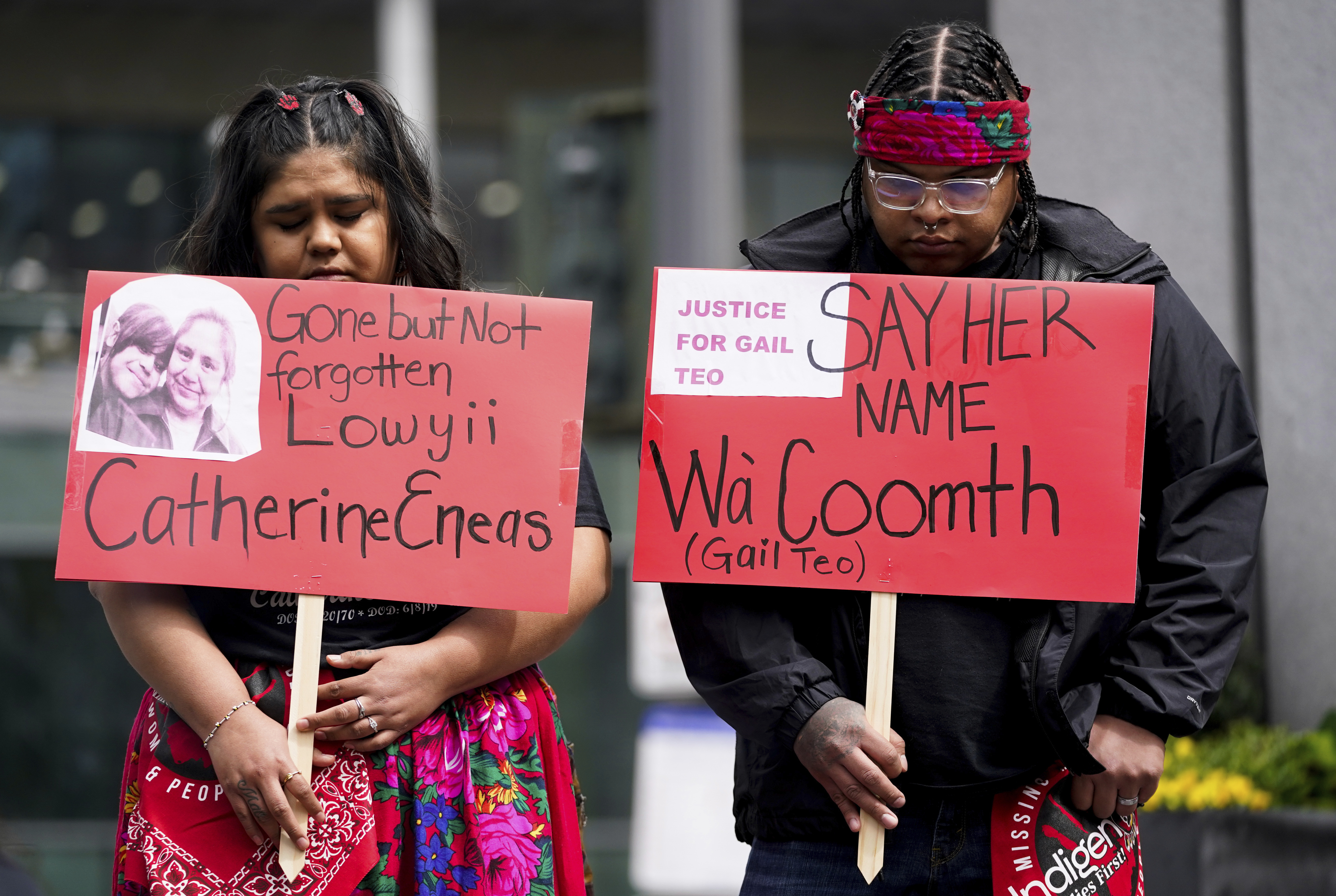 Two Native American protestors hold red signs with the names of Indigenous victims.
