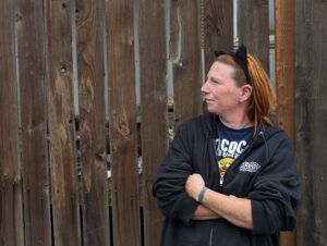 Crystal Riggs has red hair and wears black cat ears as she stands near a brown fence.
