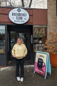 Dalia Dempster stands in front of a sign that reads "Breakfast Club" in a bright yellow sweatshirt. 