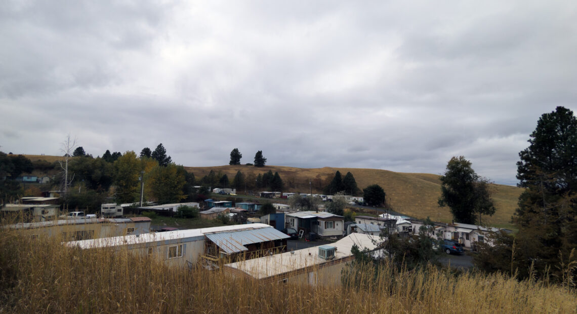 A mobile home community filled with grey and white trailer houses can be seen surrounded by tall golden grass under a cloudy sky.