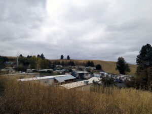 A mobile home community filled with grey and white trailer houses can be seen surrounded by tall golden grass under a cloudy sky.