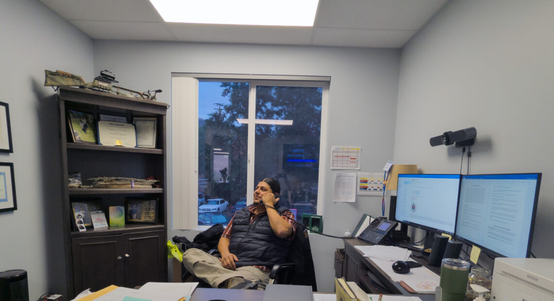 Leotis McCormack sits at his desk with double computer monitors and a brown shelf behind him, filled with knick-knacks. He holds a phone in his hand.