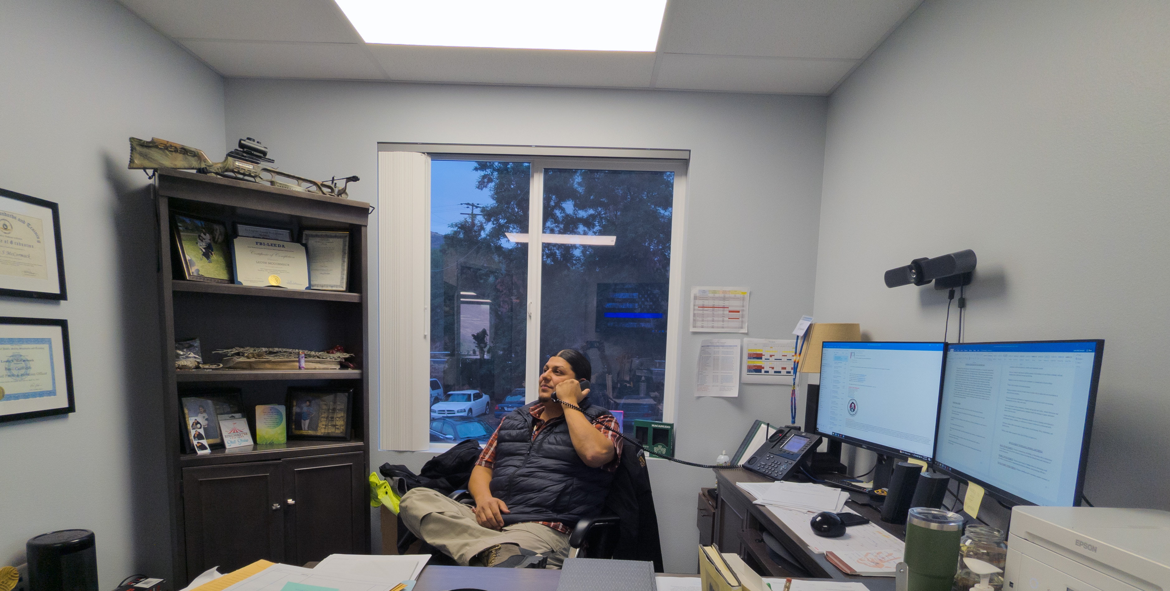 Leotis McCormack sits at his desk with double computer monitors and a brown shelf behind him, filled with knick-knacks. He holds a phone in his hand.