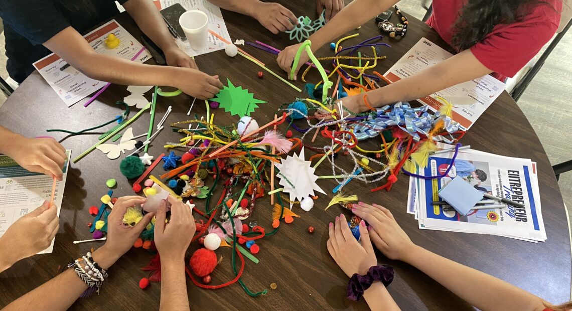 Hands of multiple teenagers rest upon a brown table as teens use pipe cleaners and pom poms alongside documents.