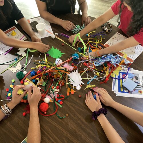 Hands of multiple teenagers rest upon a brown table as teens use pipe cleaners and pom poms alongside documents.
