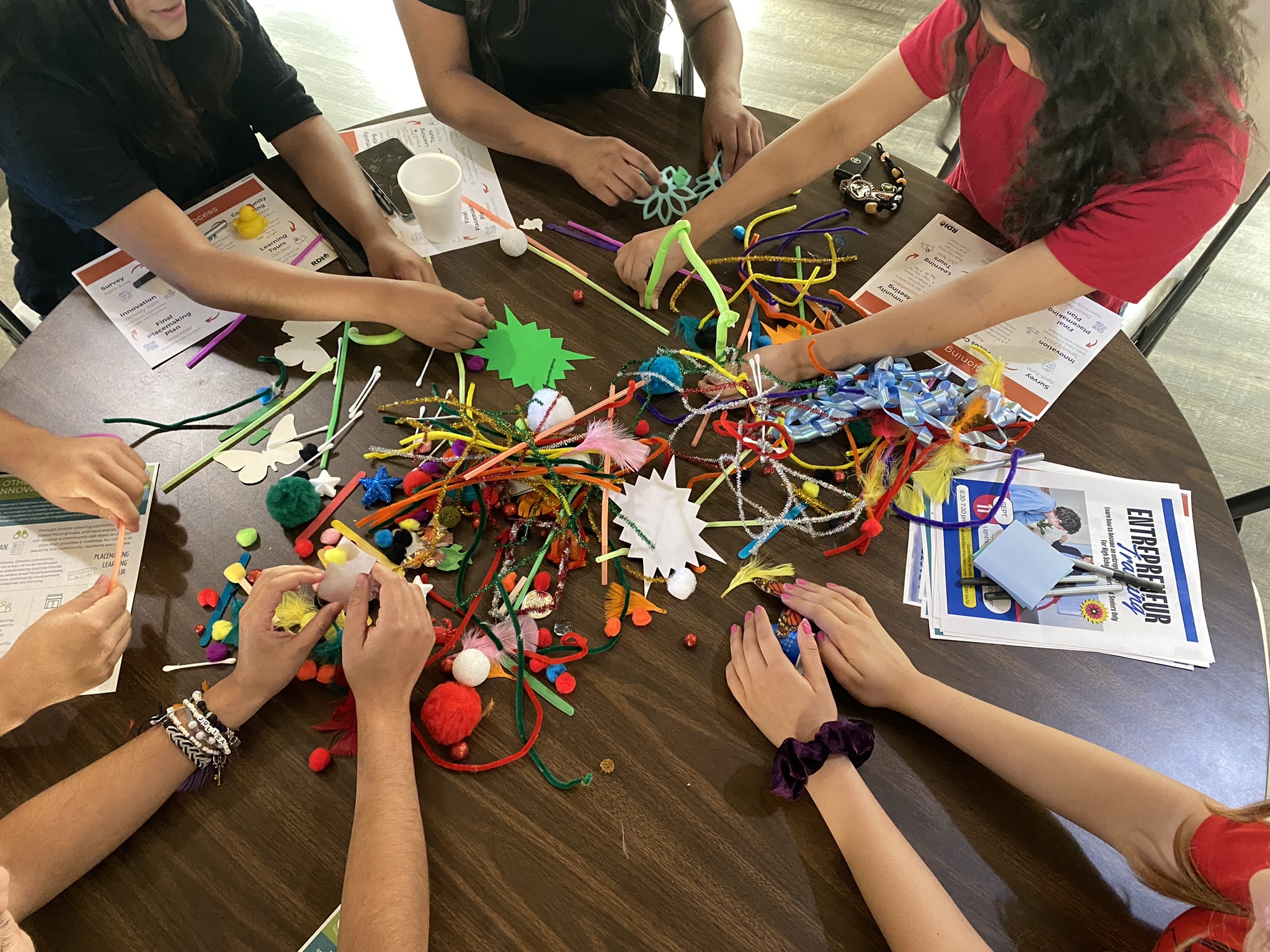 Hands of multiple teenagers rest upon a brown table as teens use pipe cleaners and pom poms alongside documents.