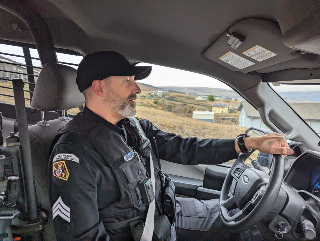 Photo of Patrol Sergeant John Svancara in uniform driving a patrol car. Grassy hills can be seen out the window. 
