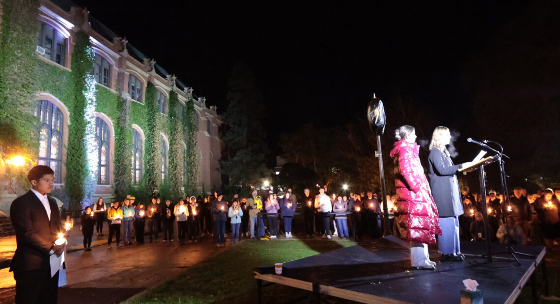 Students holding candles stand near a brick building covered in ivy illuminated by lights. A woman in a black jacket speaks on a stage before the crowd.