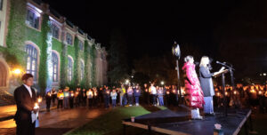 Students holding candles stand near a brick building covered in ivy illuminated by lights. A woman in a black jacket speaks on a stage before the crowd.