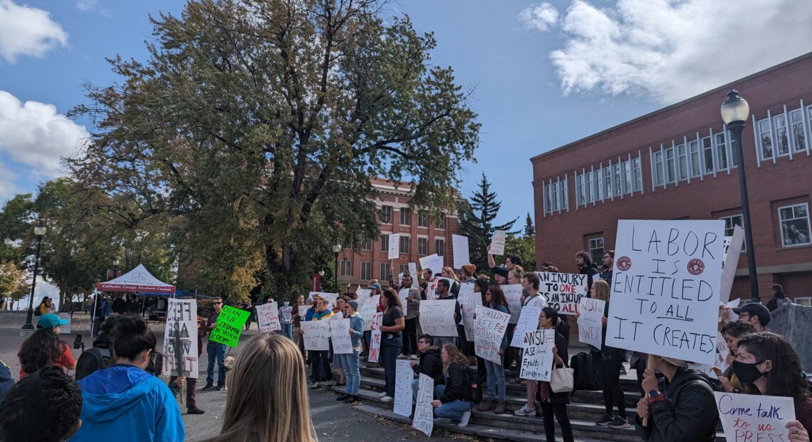 Unionized Washington State University student workers hold signs at a rally on the WSU campus. They are standing in front of a brick building next to a big tree.