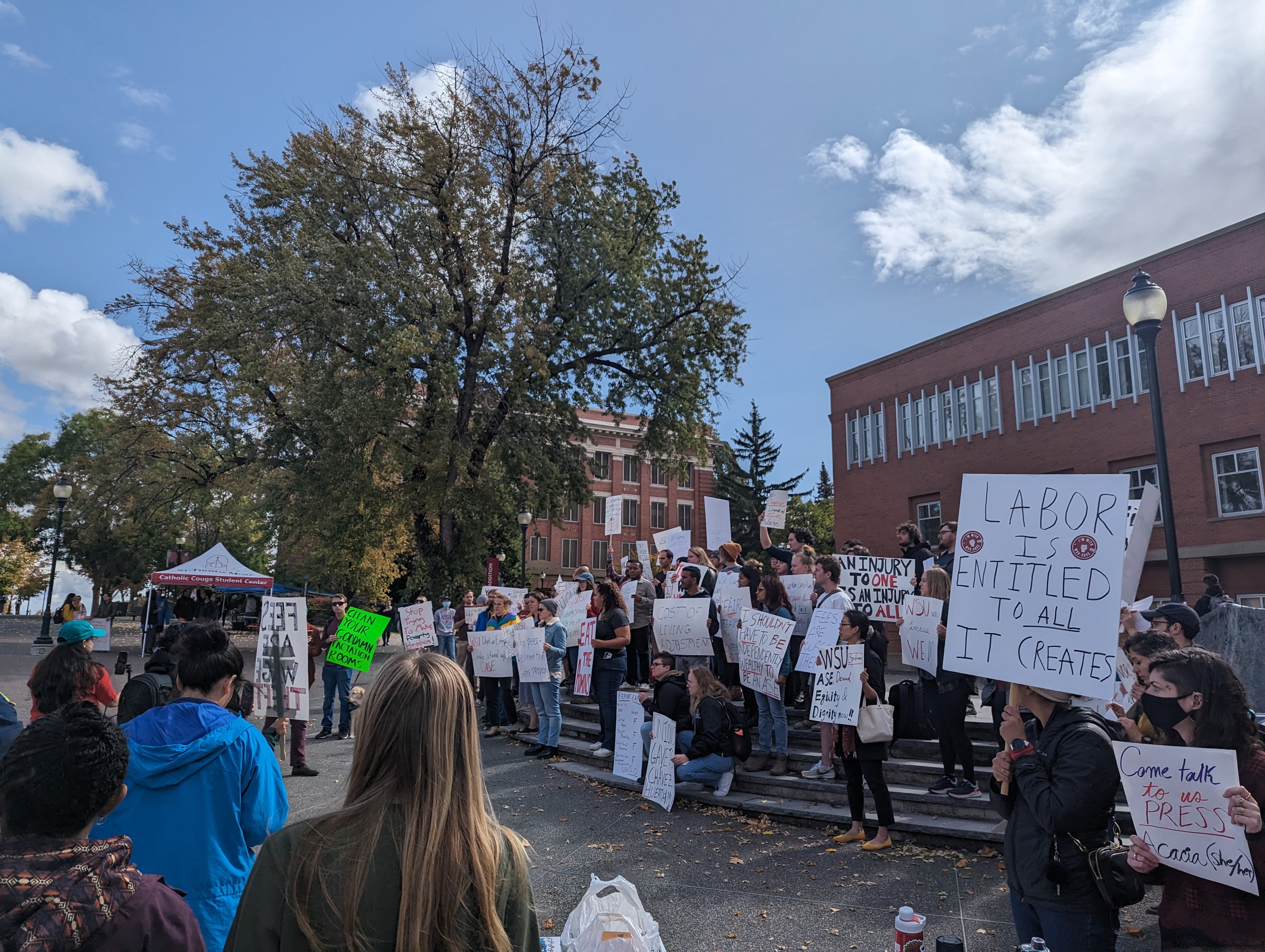 Unionized Washington State University student workers hold signs at a rally on the WSU campus. They are standing in front of a brick building next to a big tree.