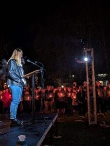 A woman with blonde hair, jeans and a black leather jacket speaks to a crowd of people holding candles.