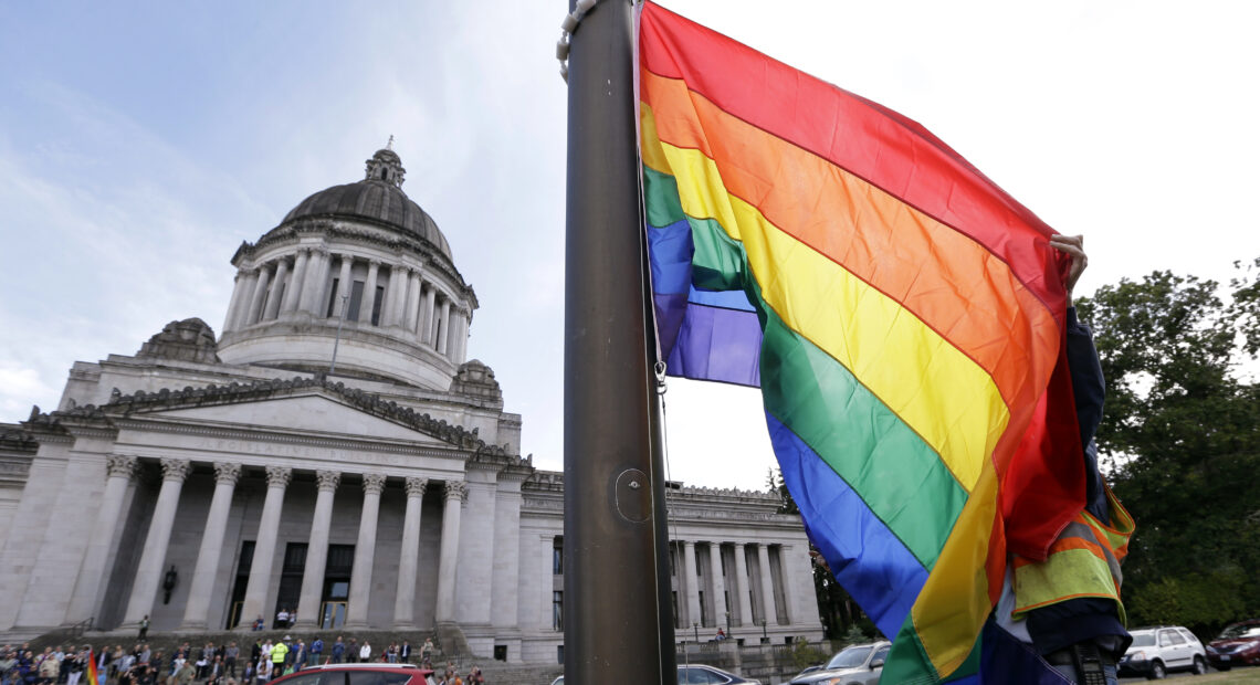 A state worker unfurls a rainbow flag in front of the Washington state Capitol. (Courtesy: Elaine Thompson / AP Photo)