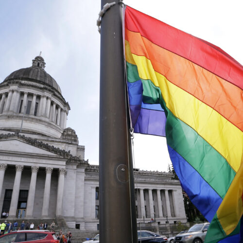 A state worker unfurls a rainbow flag in front of the Washington state Capitol. (Courtesy: Elaine Thompson / AP Photo)