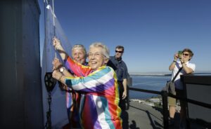 Two women with gray hair and rainbow shirts work to raise a banner against the backdrop of a blue ocean. Two men stand in the background and one is taking a picture of them with his phone.