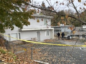 Two men in black sweatshirts stand near a large white house. A chain link fence in the front of the house has caution tape strewn across it. 