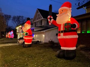 Twelve-foot-tall inflatable Santas a placed in green yards in front of white houses.