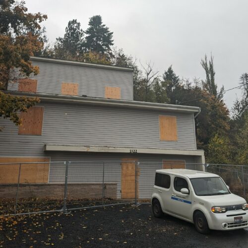 A three story dark white house with plywood in the windows sits surrounded by trees under a cloudy sky.