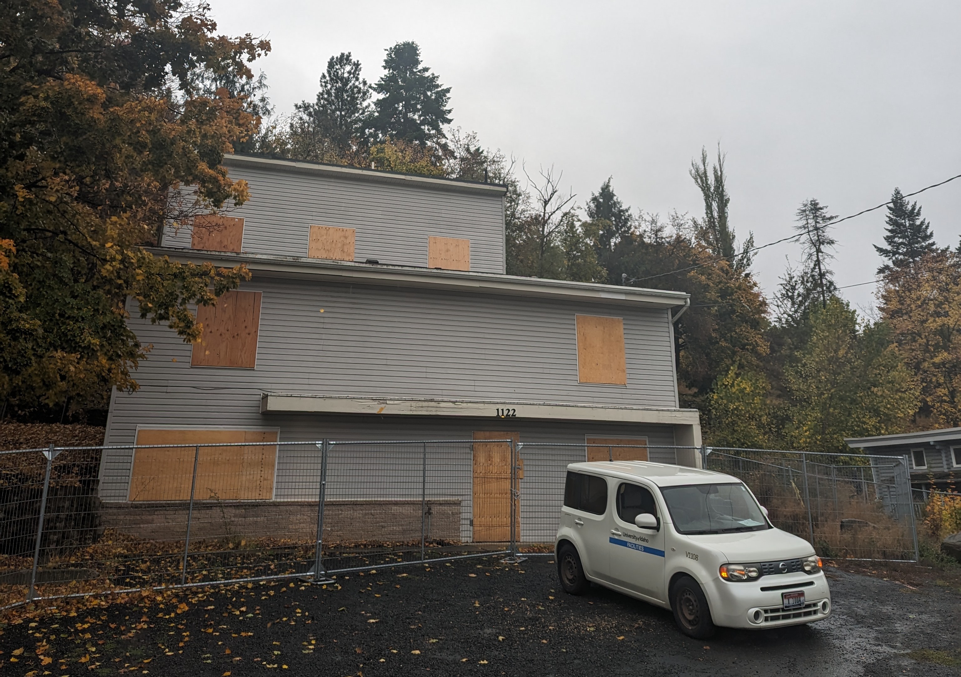 A three story dark white house with plywood in the windows sits surrounded by trees under a cloudy sky.