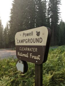 A brown and yellow sign reads "Powell Campground - Clearwater National Forest" and sits in green bushes surrounded by evergreen trees.