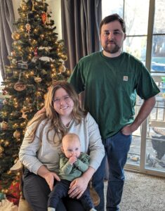 A woman in a grey sweatshirt sits in a chair with a man in a green shirt behind her. A baby sits on the woman's lap and the family poses in front of a decorated Christmas tree.