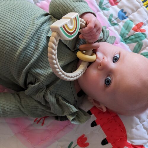 A baby in a green top holds a beige rattle toy to her month while laying on a pink blanket.