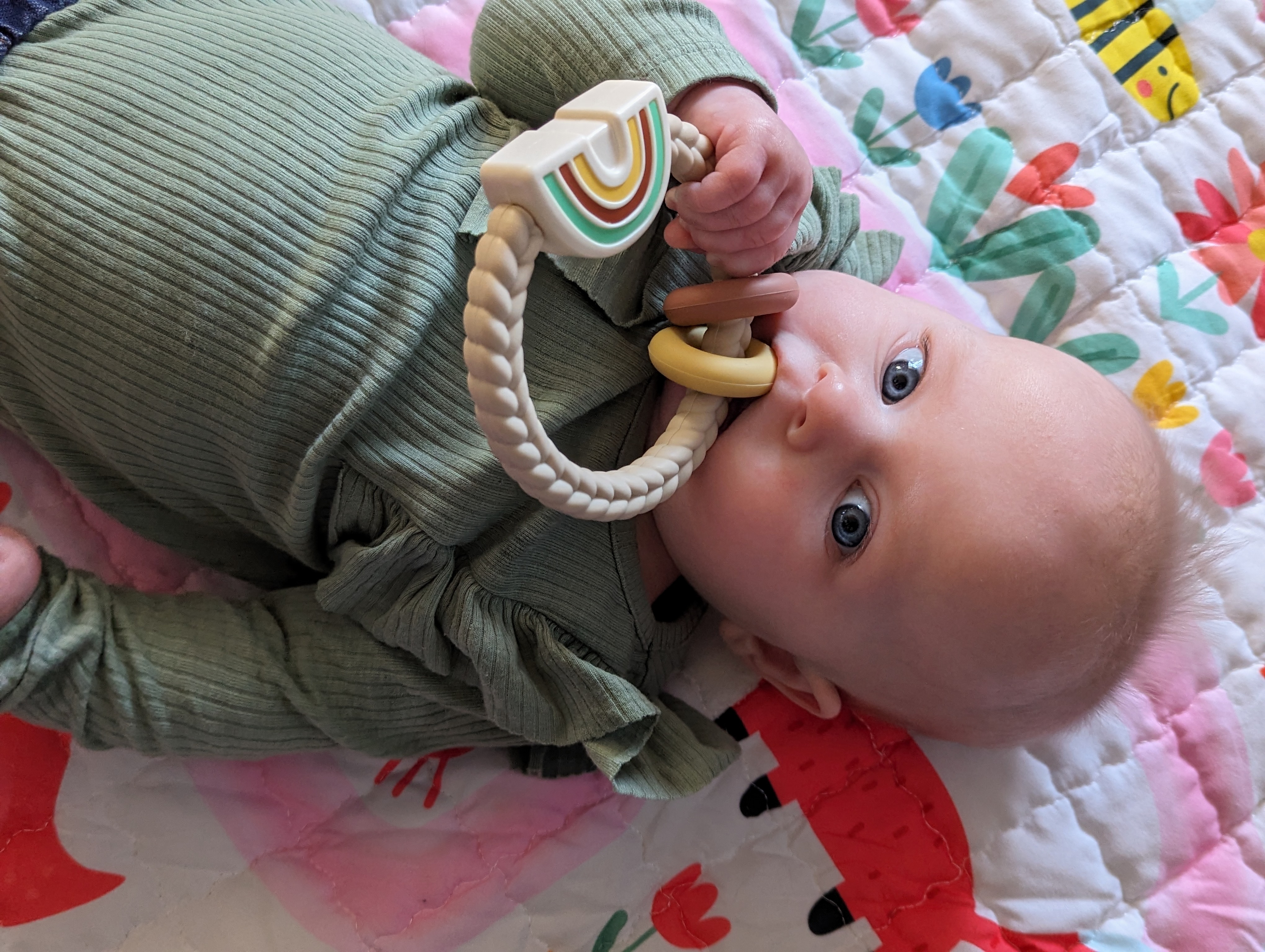 A baby in a green top holds a beige rattle toy to her month while laying on a pink blanket.