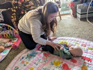 A woman in a grey sweatshirt and glasses tickles her baby who lays on a pink blanket.