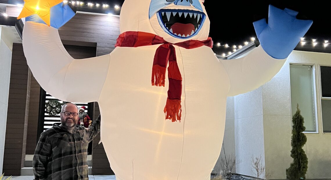 A man in a plaid shirt with grey pants on stands next to a 12-foot-tall white and blue inflatable Yeti.