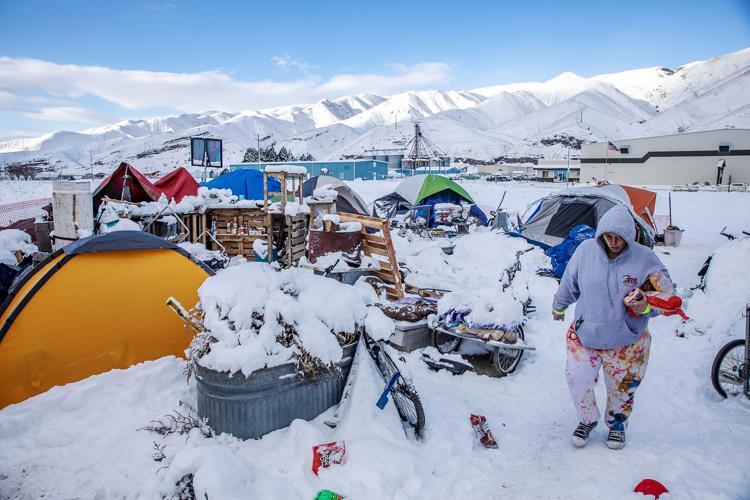 A woman walks in snow at a houseless camp with snowy hills in the background.