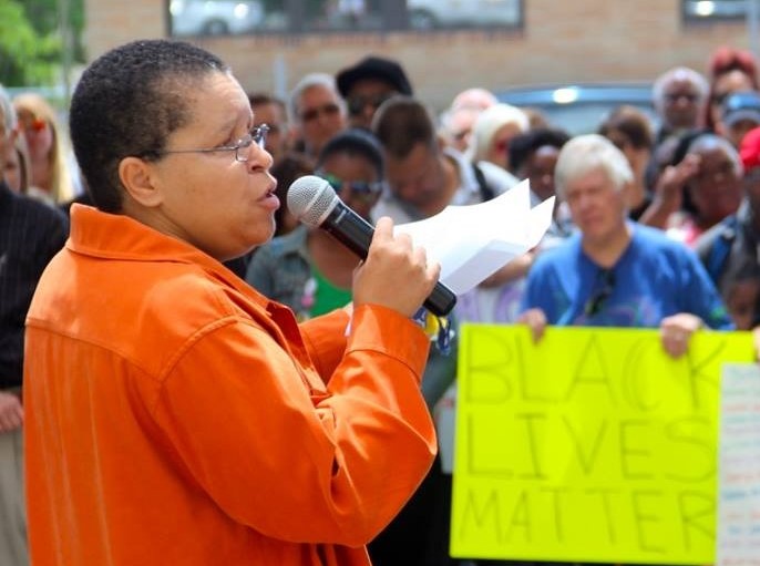 A woman in an orange shirt speaks into a microphone at a Black Lives Matter rally.
