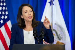 Senator Cantwell stands at a podium in a blue blazer with an American flag behind her.